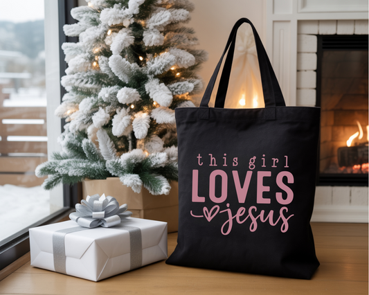 Black tote bag with 'this girl loves Jesus' text in pink, placed in front of a decorated Christmas tree and fireplace.