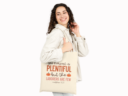 Smiling woman in a light jacket holds a beige tote bag over her shoulder. The bag reads "The harvest is plentiful but the laborers are few, Matthew 9:37" with pumpkin graphics.