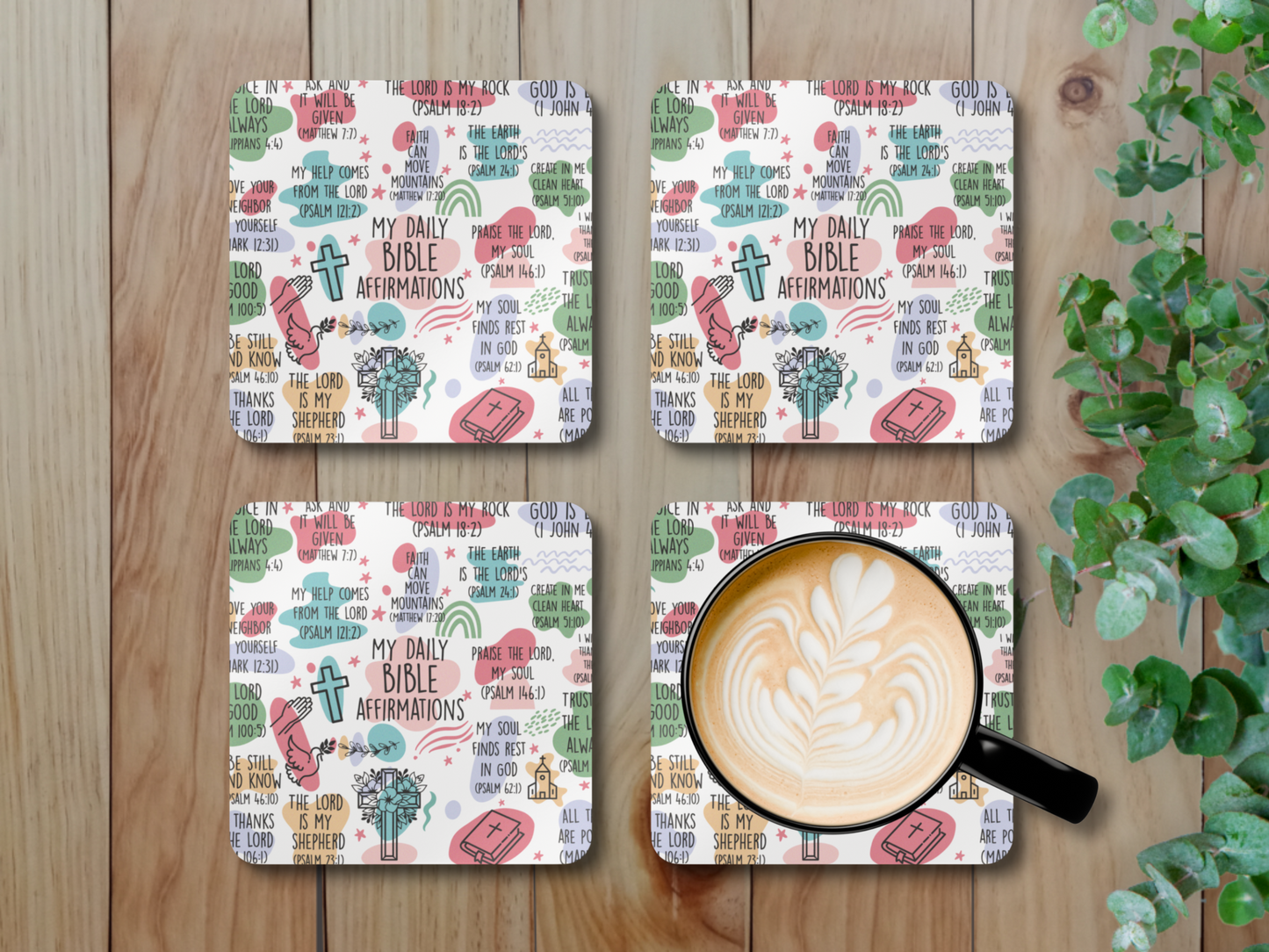 Set of four colorful scripture affirmation coasters with latte art coffee mug, displayed on wooden surface with green leafy plant.