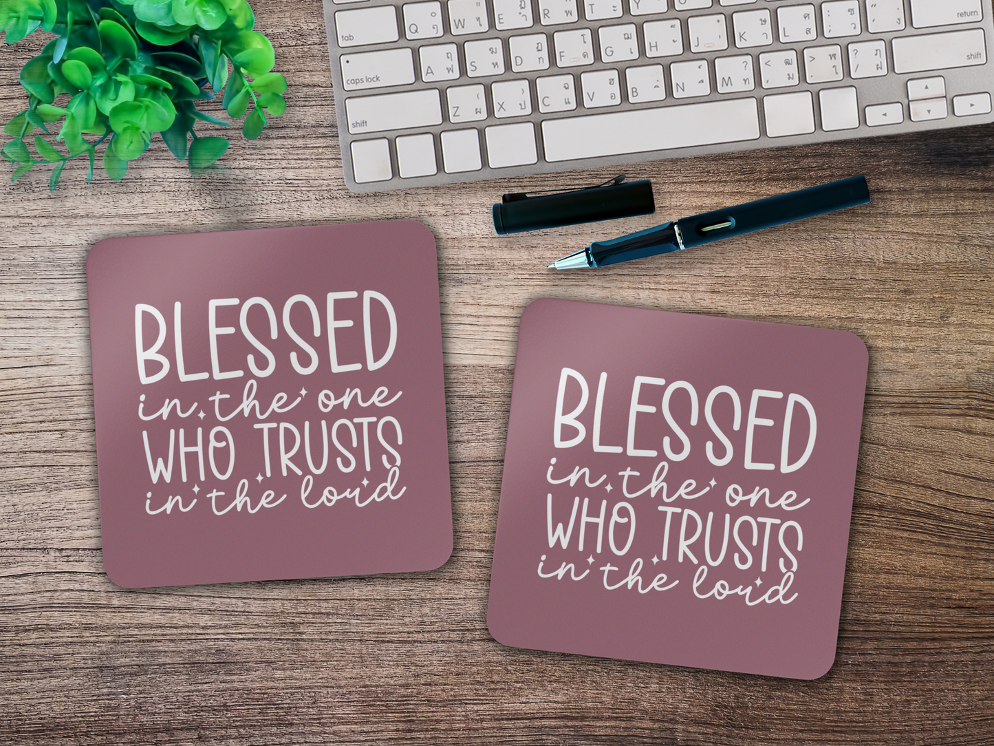 Two mauve scripture coasters on a wooden desk beside a keyboard, pen, and plant.