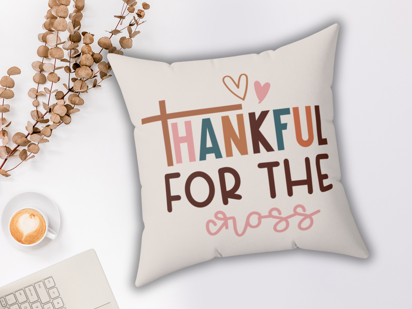 A decorative pillow with the text "Thankful for the Cross" in colorful letters. A cappuccino with a heart design and dried eucalyptus are nearby, creating a cozy atmosphere.