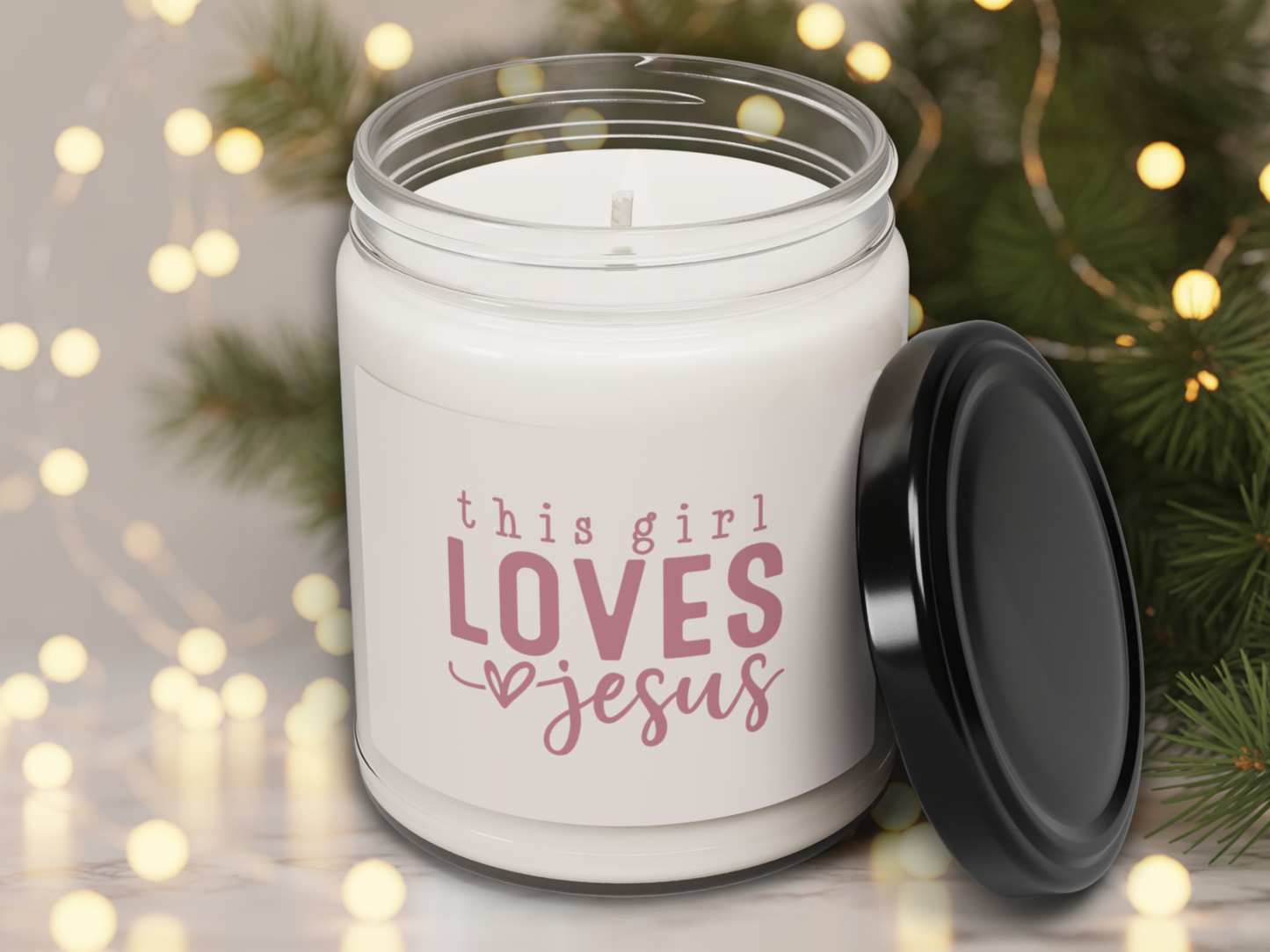 White candle jar with 'this girl loves Jesus' text, black lid, against a festive background with lights and greenery.