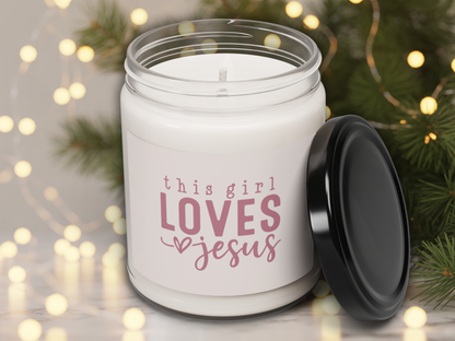 White candle jar with 'this girl loves Jesus' text, black lid, against a festive background with lights and greenery.