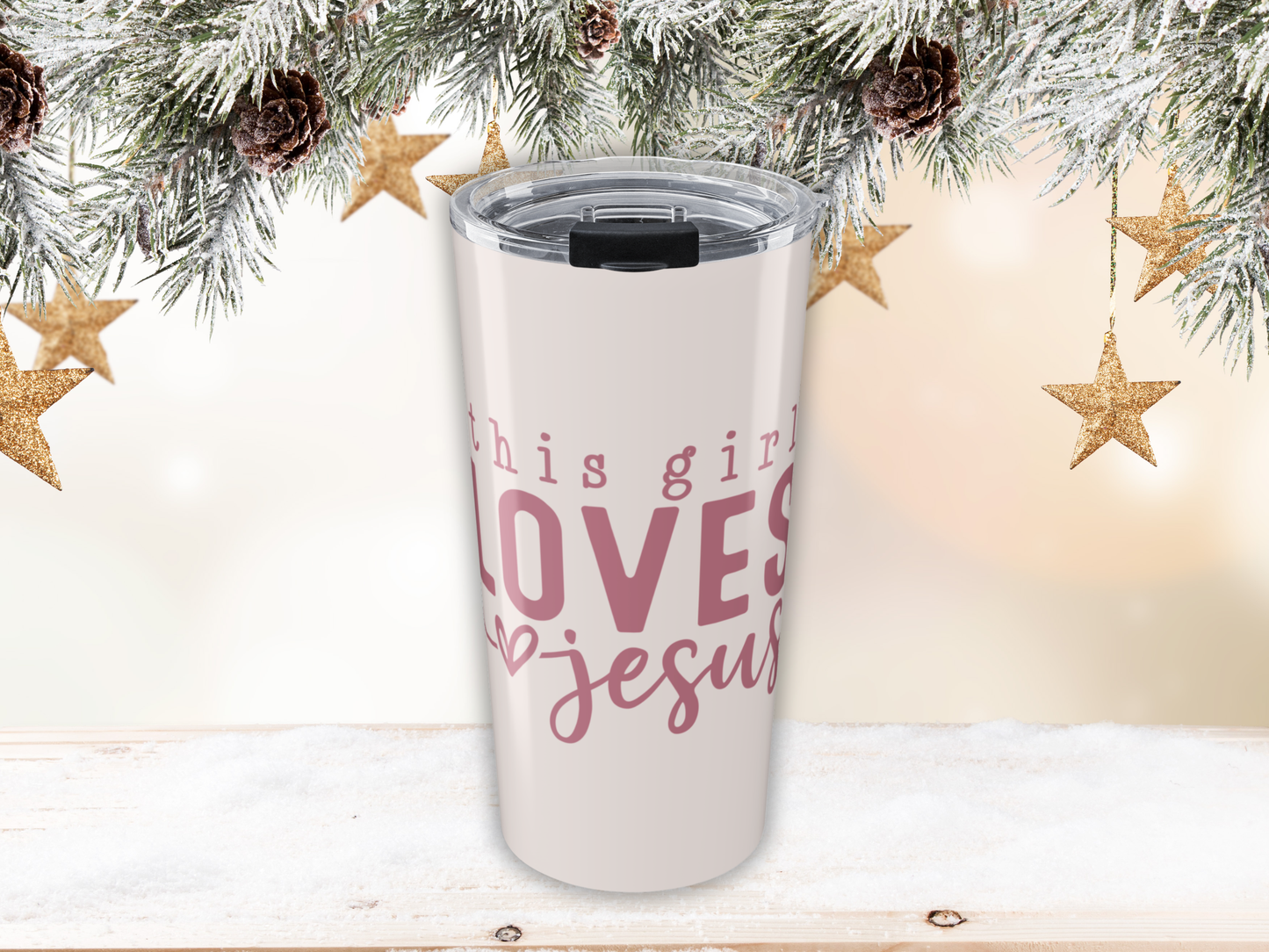 Pink tumbler with pink text 'This girl loves Jesus' against a festive background with stars and pine cones.