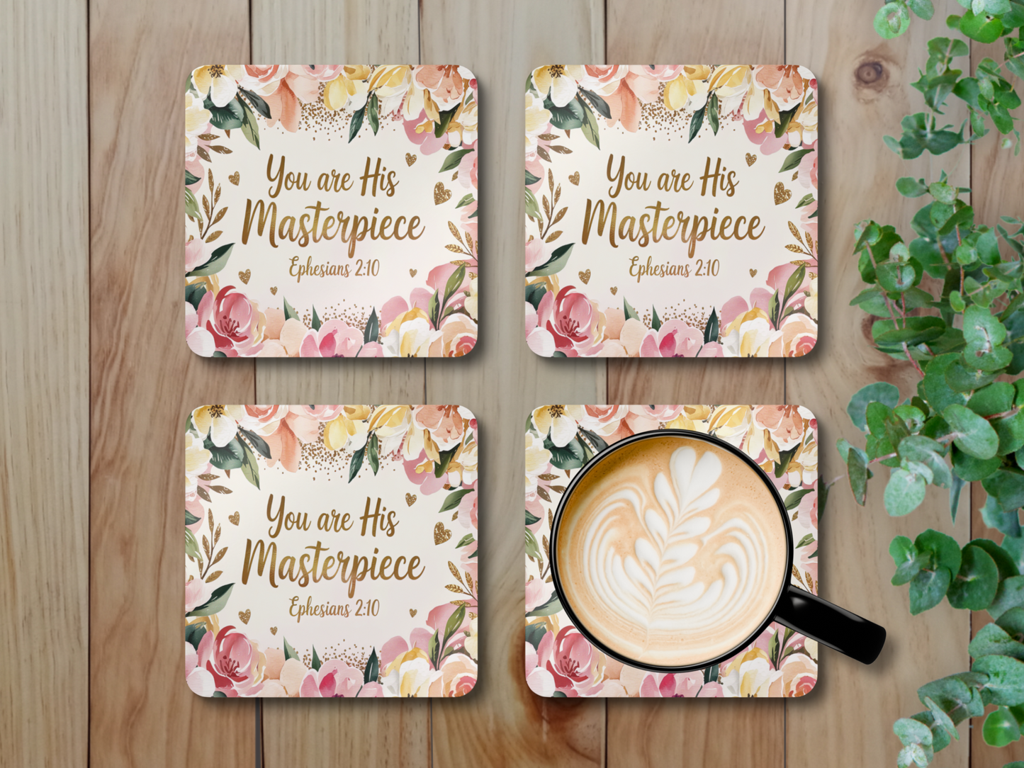 Set of four scripture coasters with latte art coffee mug on one, displayed on wooden surface beside green leafy plant.