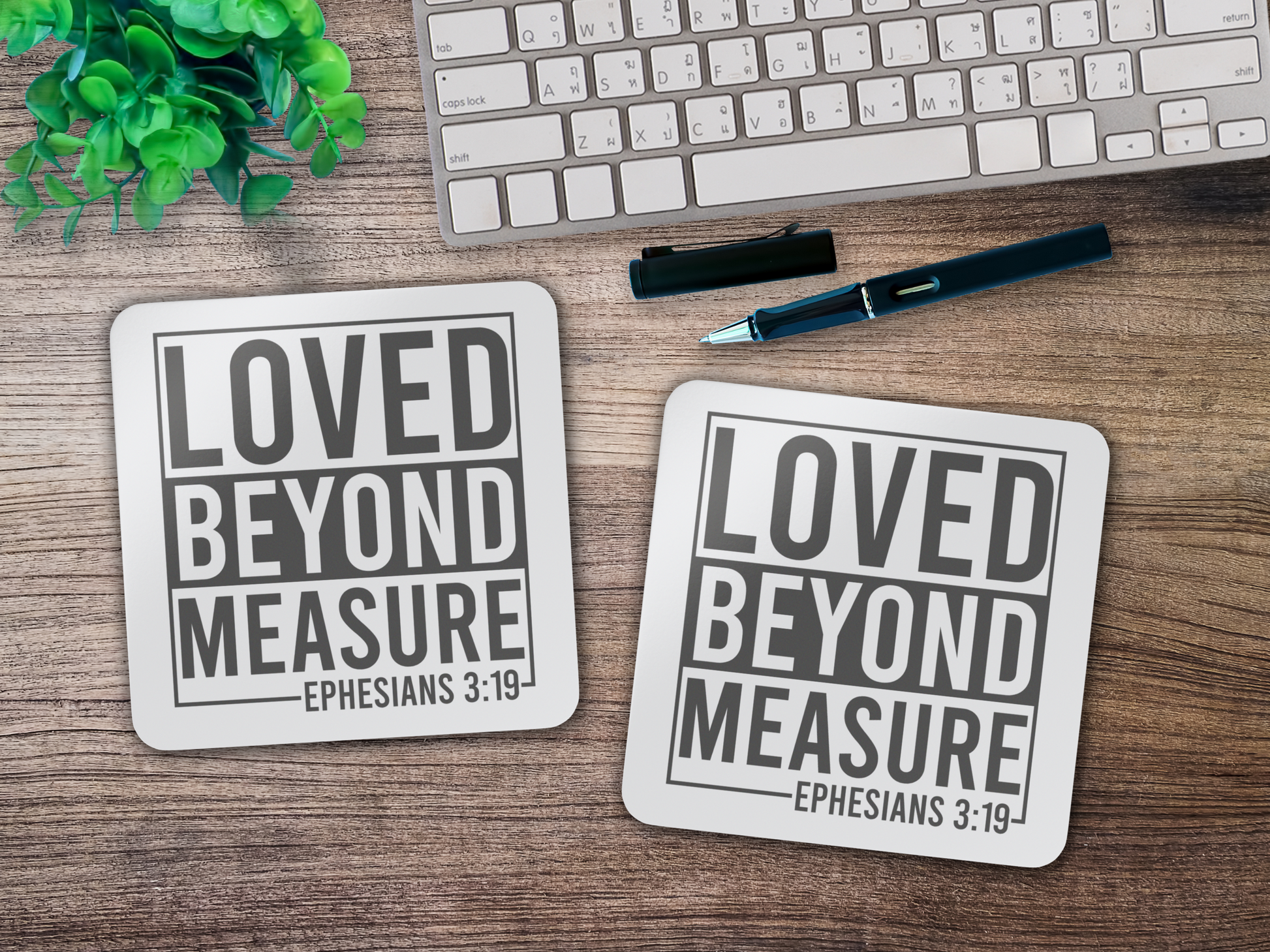 Two scripture coasters on a wooden desk beside a keyboard, pen, and potted plant.