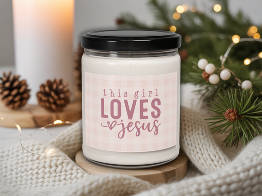 Candle jar with 'this girl LOVES Jesus' text on a cozy background with lights and pine cones.