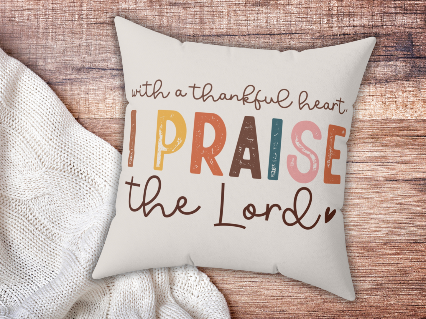 Cushion on wooden surface with text: "With a thankful heart, I praise the Lord" in multicolored, playful fonts. Cozy and uplifting mood.