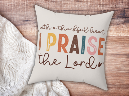 Cushion on wooden surface with text: "With a thankful heart, I praise the Lord" in multicolored, playful fonts. Cozy and uplifting mood.