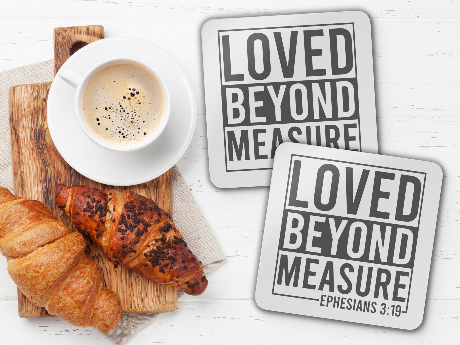 Two “Loved Beyond Measure” coasters beside coffee and croissants on a wooden board, white tabletop setting.