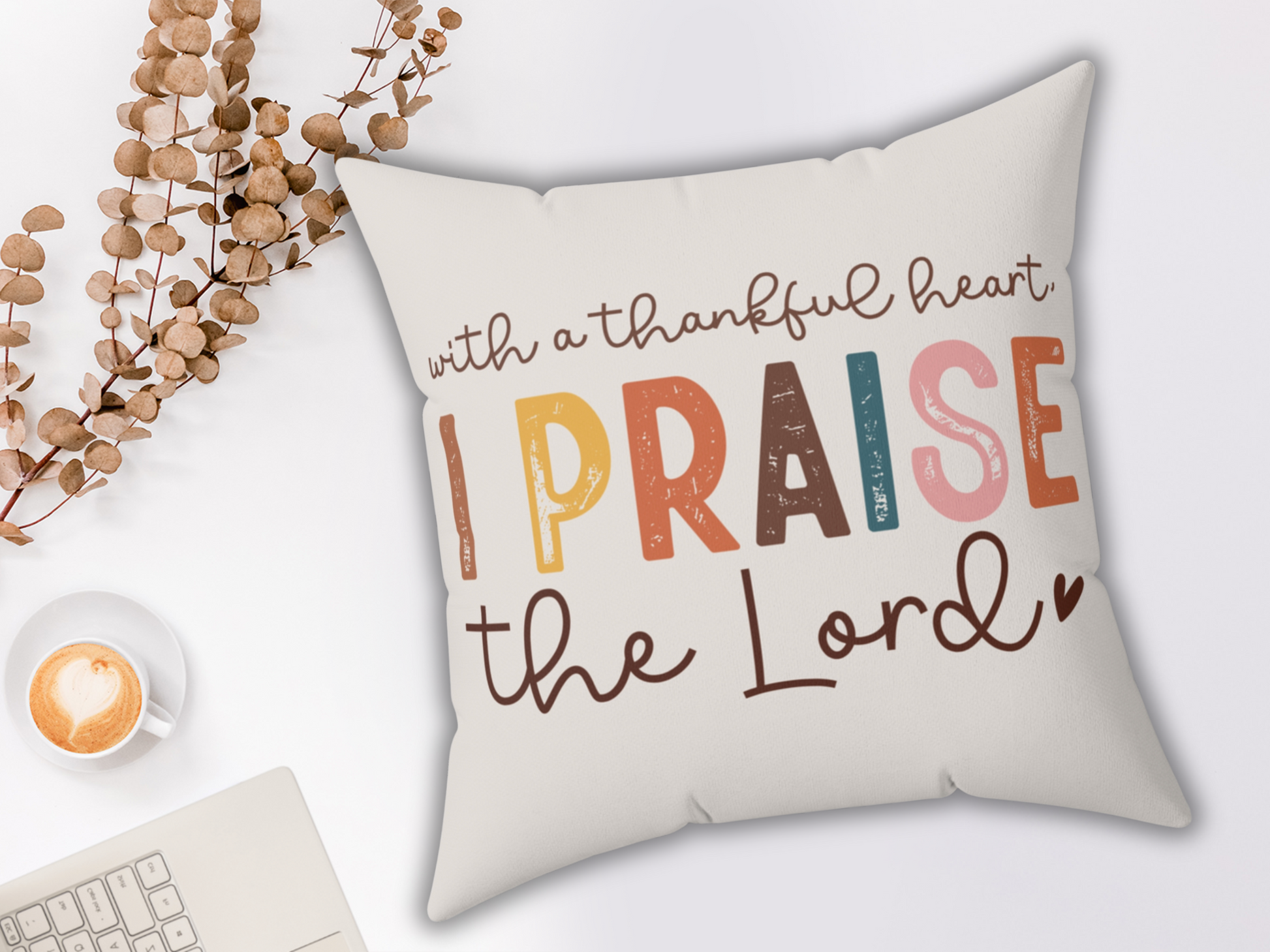 A pillow with colorful text that reads "With a thankful heart, I praise the Lord." Nearby are dried leaves, a latte with a heart in the foam, and a laptop.