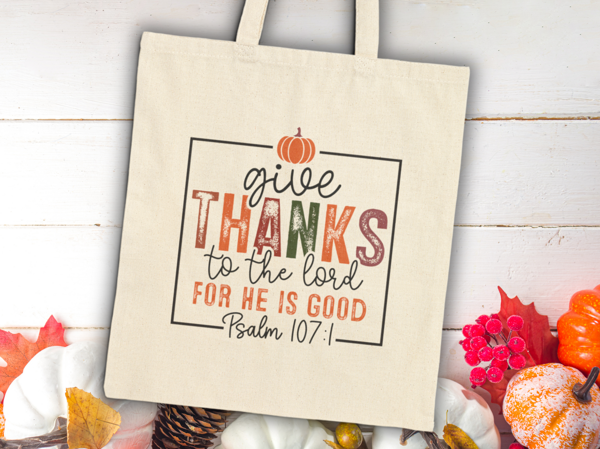 Canvas tote bag on whitewashed wood, featuring "Give Thanks" text with a pumpkin graphic. Surrounded by pumpkins and leaves, evoking a festive, grateful tone.