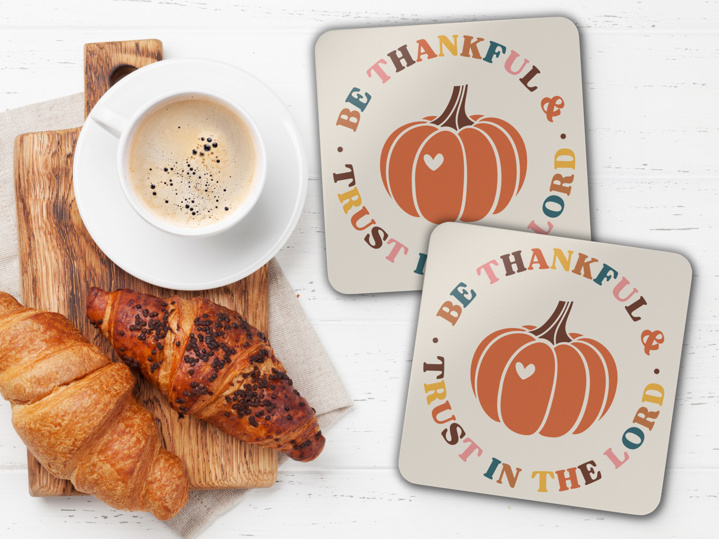 Pair of scripture pumpkin coasters with coffee and croissants on wooden serving board over white tabletop.