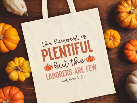 Cream tote bag on a wooden surface surrounded by various pumpkins. Text reads "The harvest is plentiful but the laborers are few, Matthew 9:37."