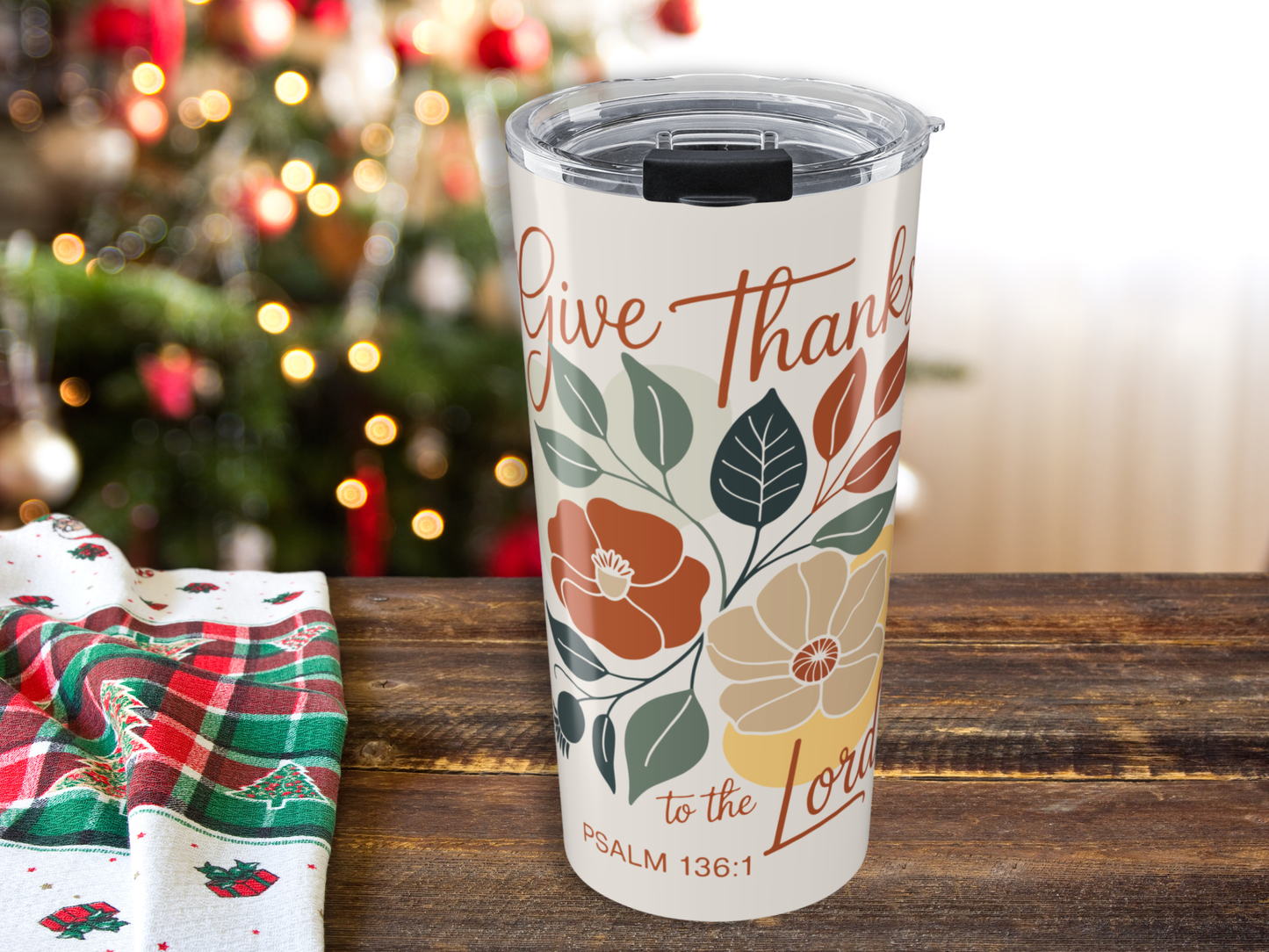 Tumbler with 'Give Thanks to the Lord' design on a wooden table with a Christmas tree in the background.