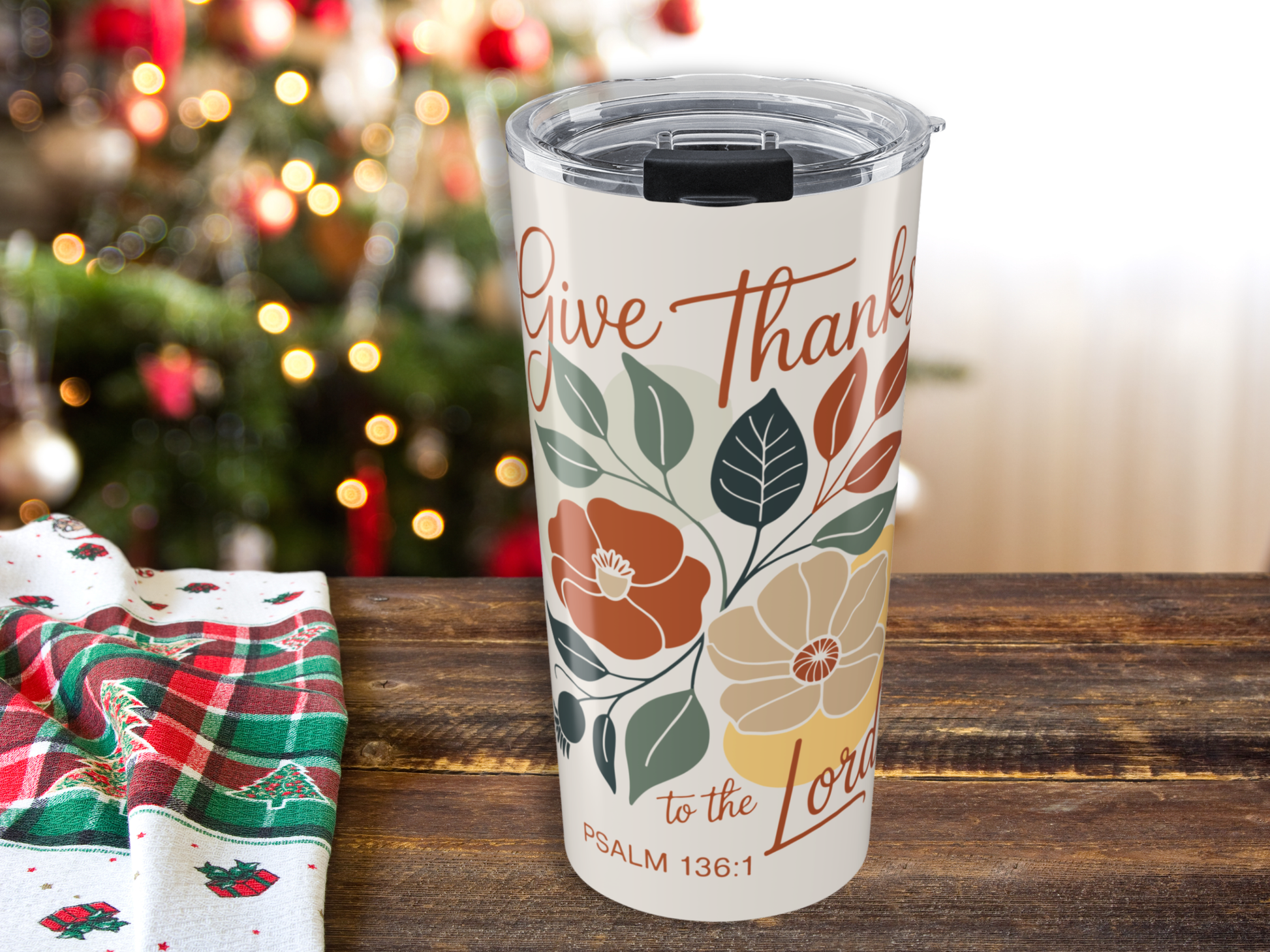 Tumbler with 'Give Thanks to the Lord' design on a wooden table with a Christmas tree in the background.