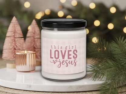 Candle jar with 'this girl loves Jesus' text on a decorative surface with Christmas trees and lights.