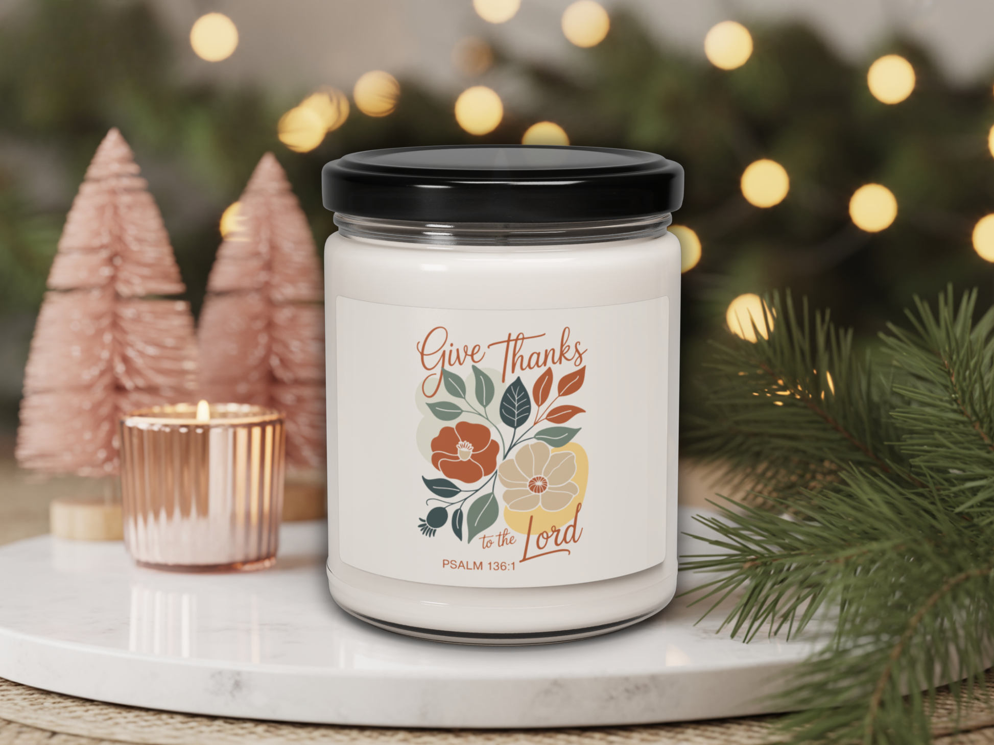 Candle jar with 'Give Thanks to the Lord' text and floral design on a decorative setting with lights and trees.
