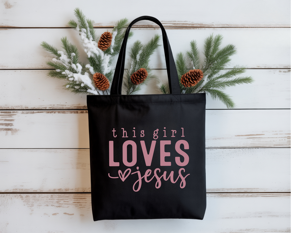 Black tote bag with 'this girl LOVES Jesus' text on a wooden surface with decorative branches.