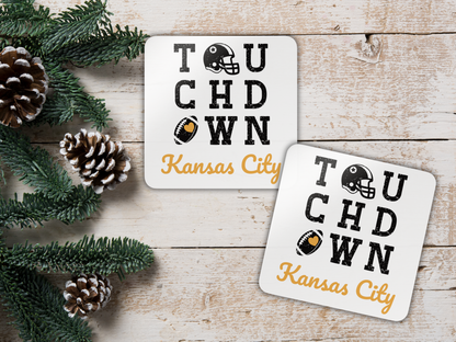 Two 'Touchdown Kansas City' coasters on a wooden surface with pine cones and greenery.
