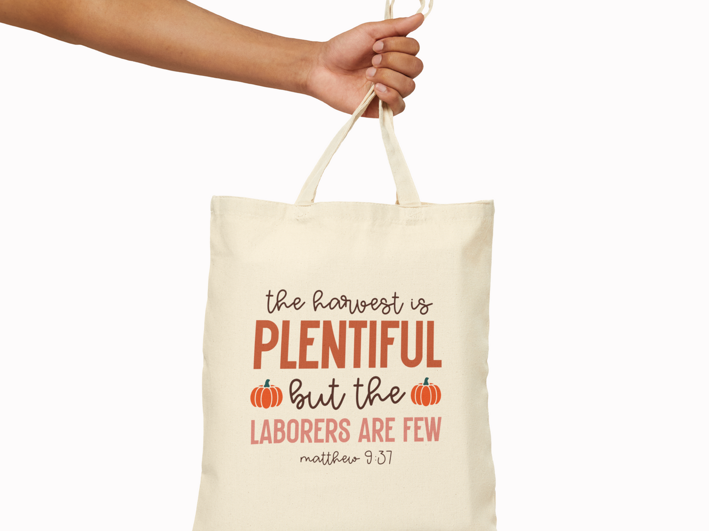 A hand holds a beige tote bag with text: "The harvest is plentiful, but the laborers are few. Matthew 9:37," decorated with small pumpkins. Tone: warm and inviting.