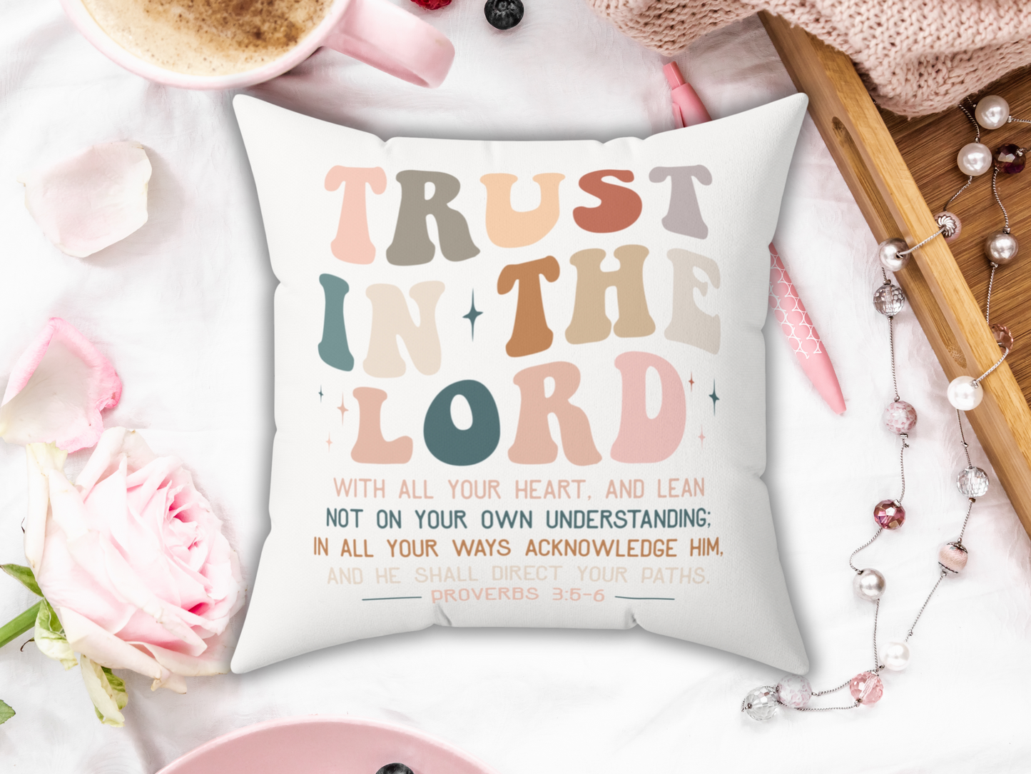 Decorative Bible verse pillow reading “Trust in the Lord,” placed on white bedding with pink rose, coffee, pen, and wooden tray.