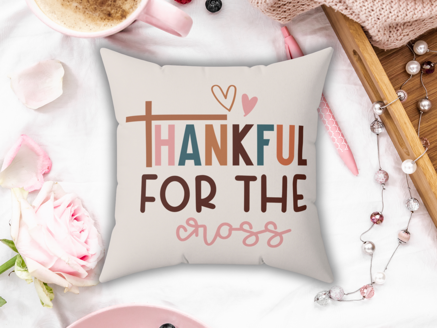 A decorative Christian pillow on a white surface reads "Thankful for the cross" with heart symbols. Surrounding items include a pink rose, cappuccino, berries, and beads, creating a warm, cozy atmosphere.