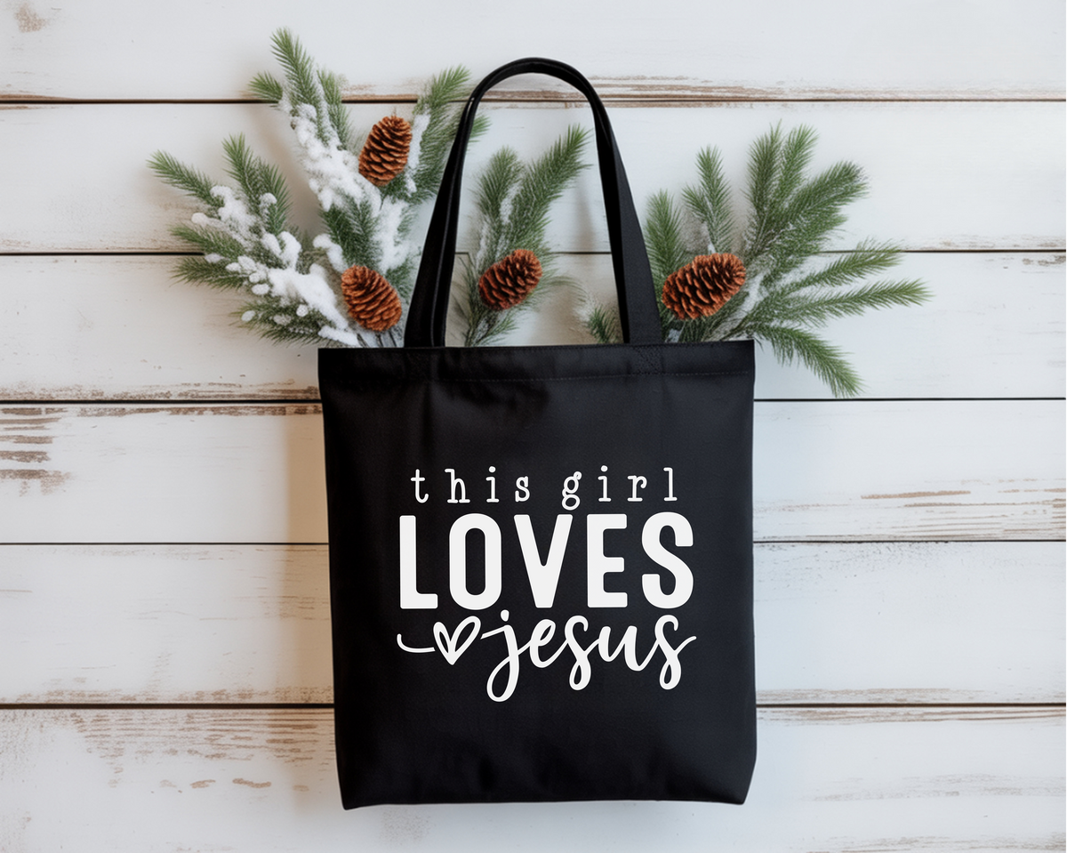 Black tote bag with 'this girl loves Jesus' text on a wooden surface with decorative branches.