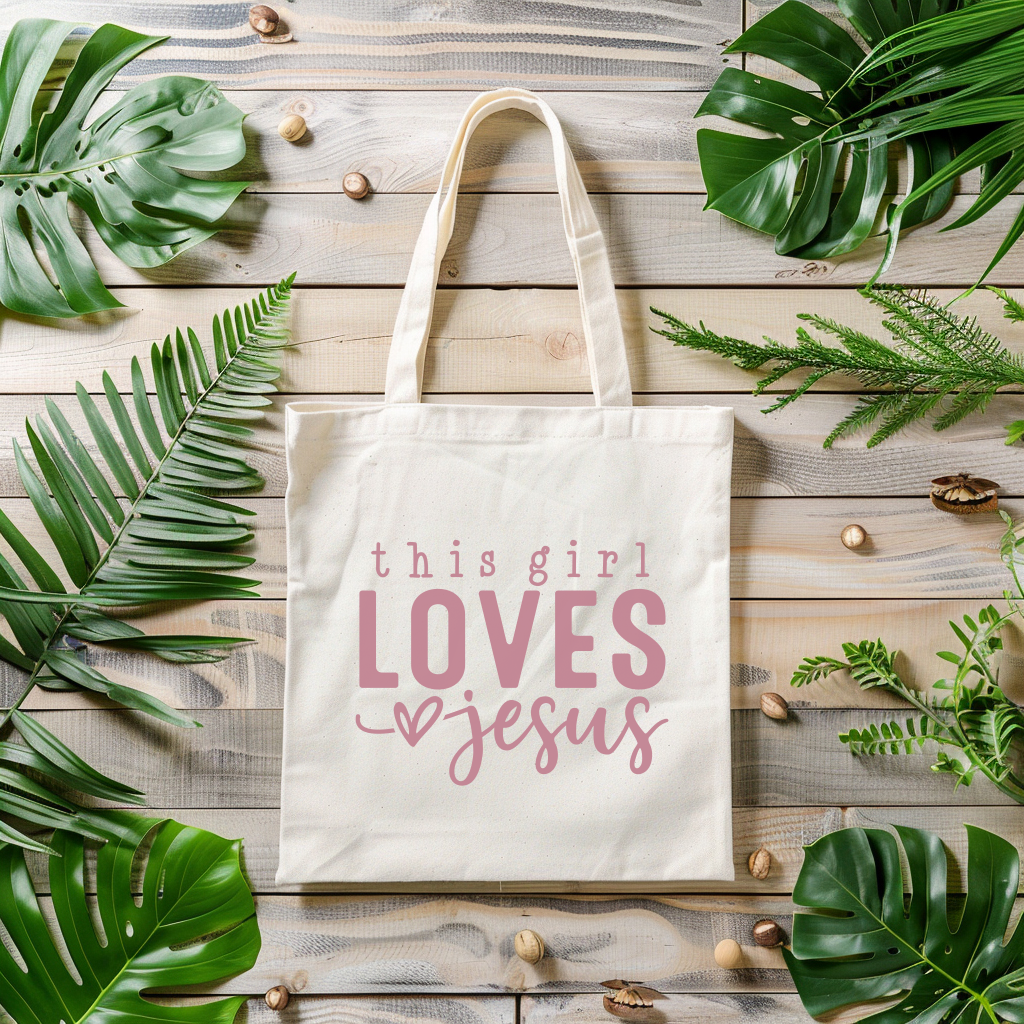 Tote bag with 'this girl LOVES Jesus' text on a wooden surface with green leaves.