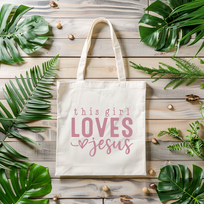 Tote bag with 'this girl LOVES Jesus' text on a wooden surface with green leaves.