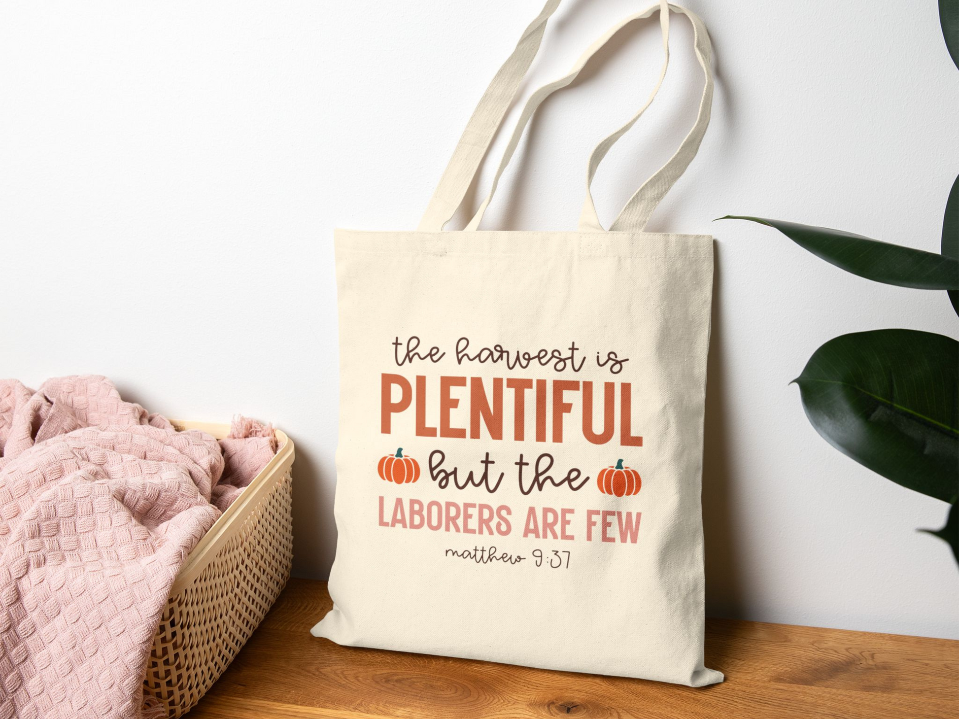 A cream tote bag rests on a wooden surface next to a basket with pink fabric. The bag displays the text "The harvest is plentiful but the laborers are few" with small pumpkin graphics, conveying a fall theme.