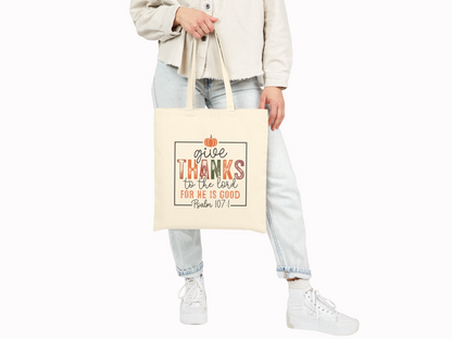 Person in a beige shirt and light jeans holding a tote bag with a fall-themed quote, "Give thanks to the Lord for He is good," decorated with a small pumpkin. Casual and warm tone.