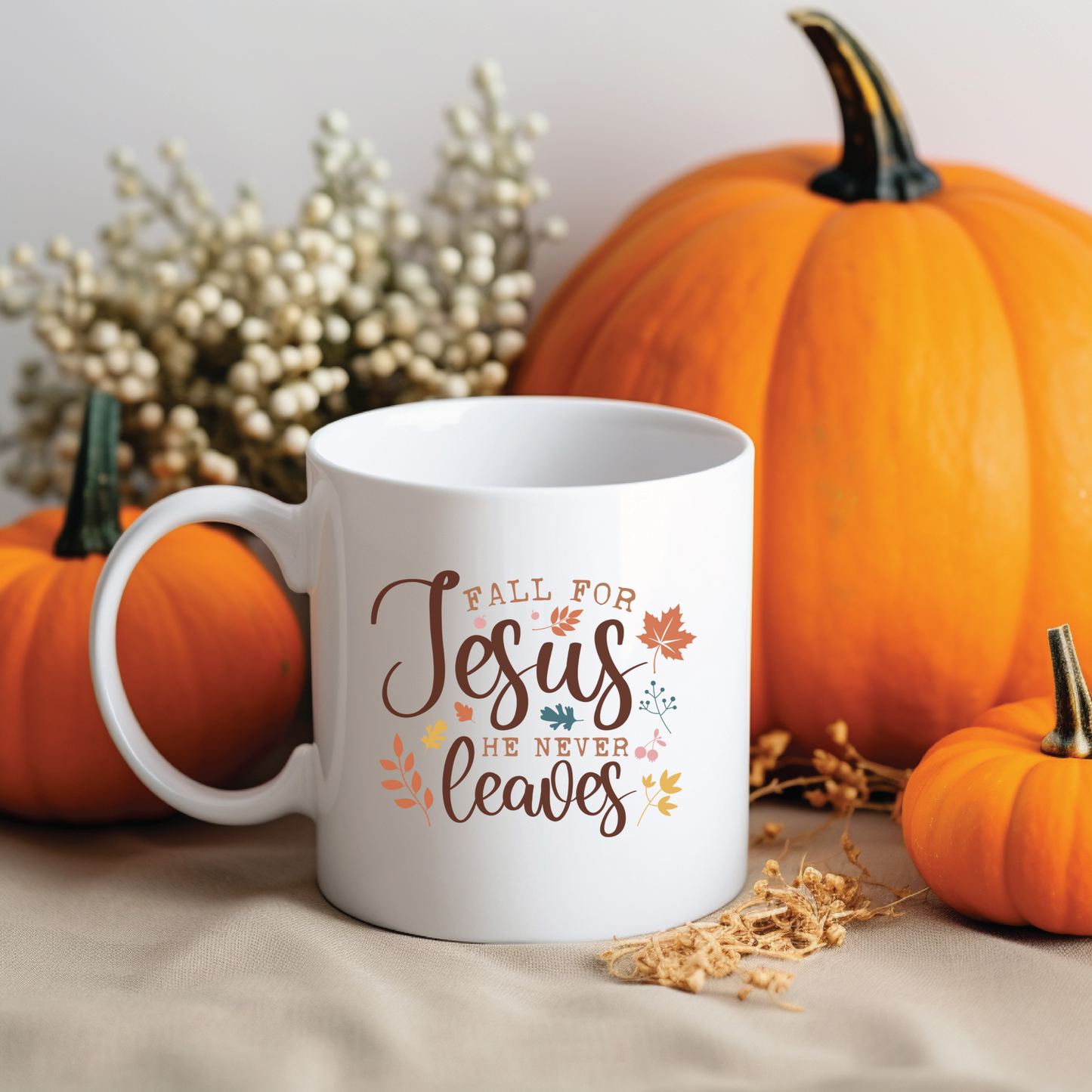 White mug with 'Fall for Jesus he never leaves' text, surrounded by pumpkins and dried foliage on a neutral background