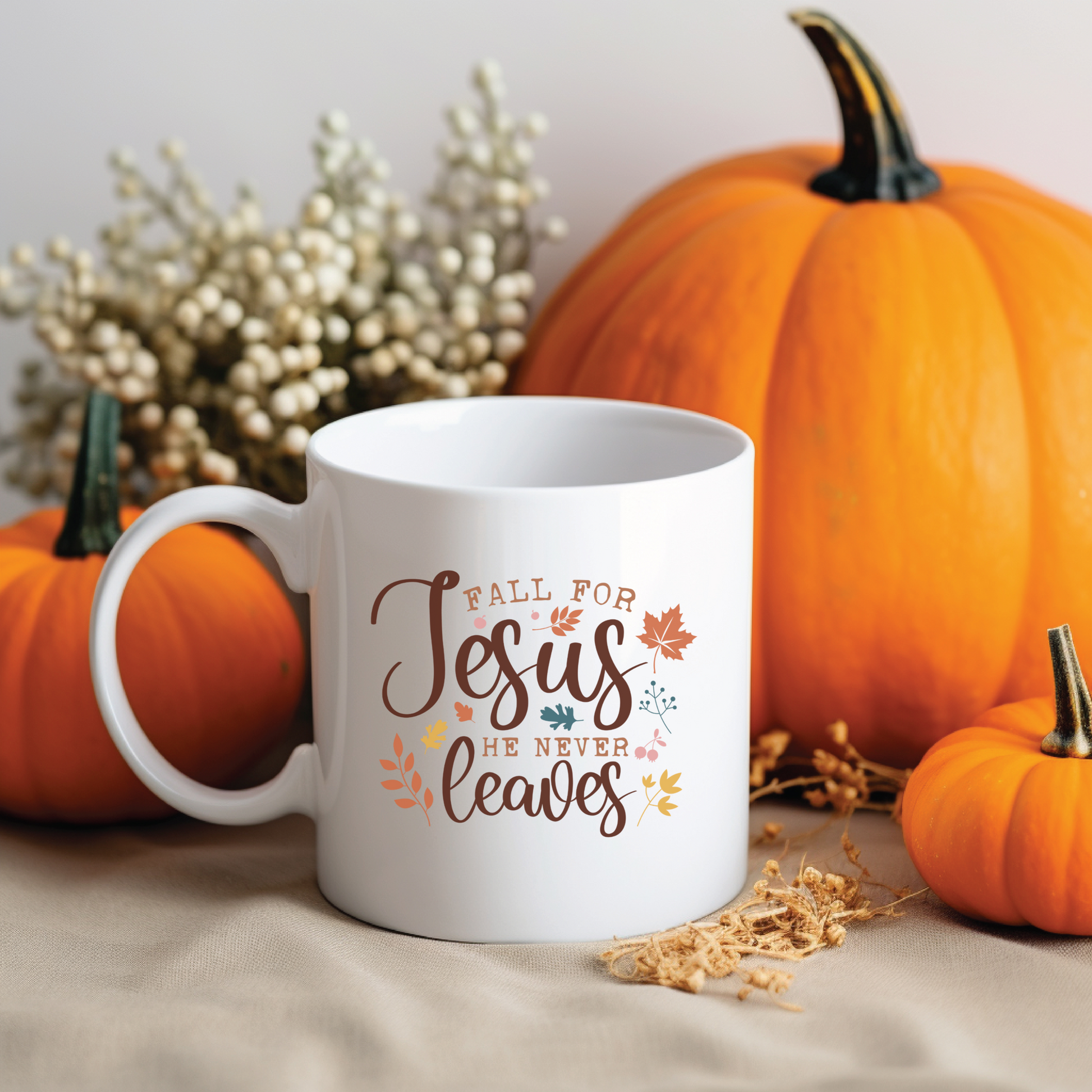 White mug with 'Fall for Jesus he never leaves' text, surrounded by pumpkins and dried foliage on a neutral background