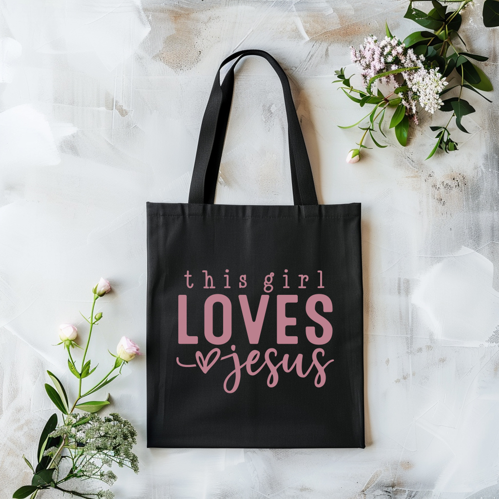 Black tote bag with 'this girl LOVES Jesus' text on a marble surface with flowers.