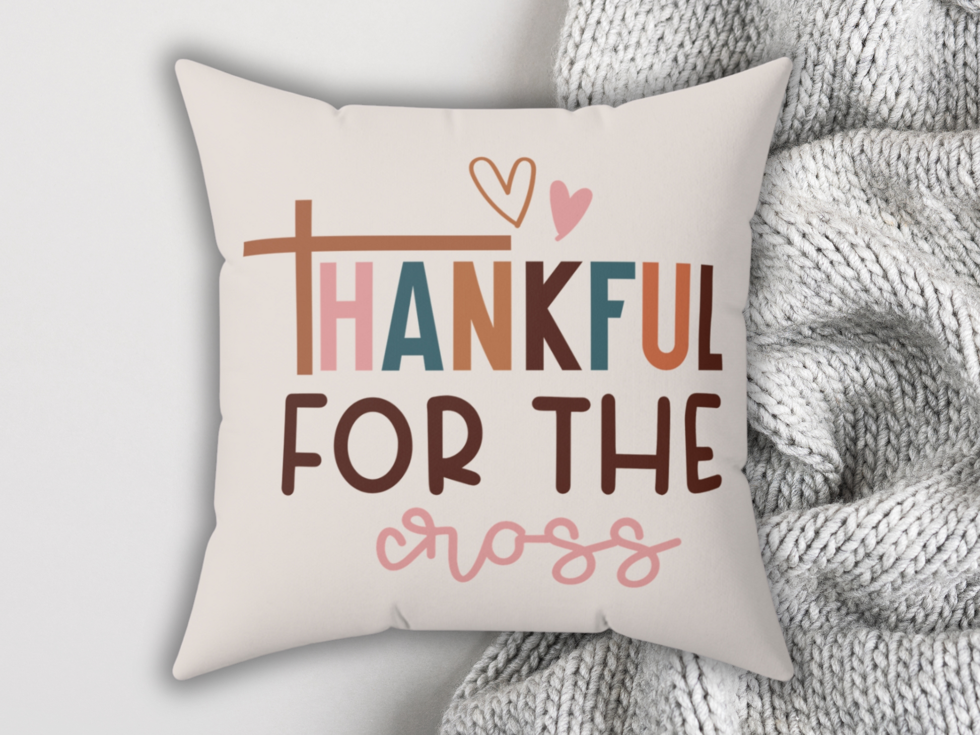 A fall pillow with the phrase "Thankful for the Cross" in colorful letters, featuring heart and cross motifs, beside a gray knitted blanket.
