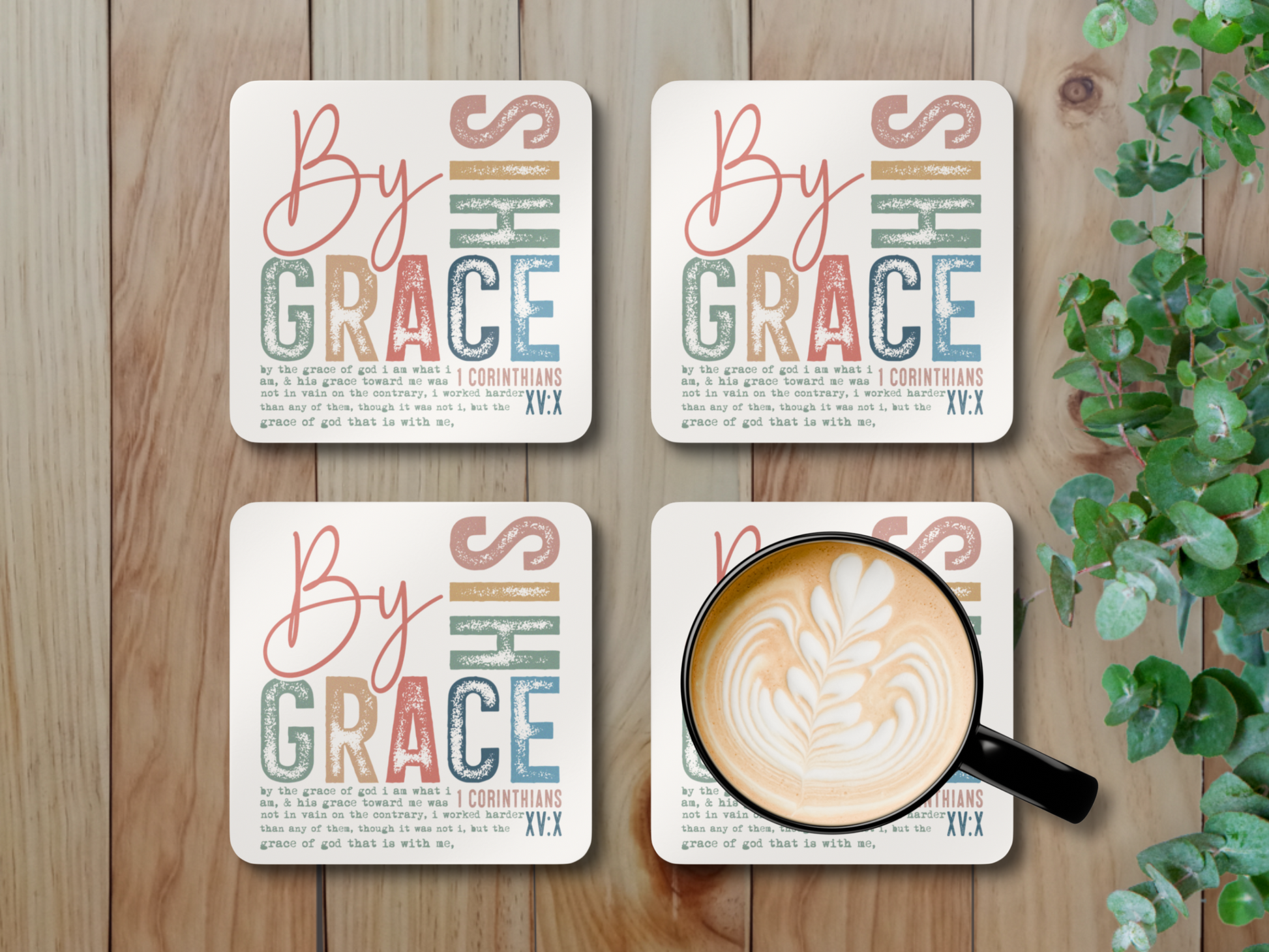 Set of four “By His Grace” coasters with latte mug styled on wooden surface beside green leafy plant.