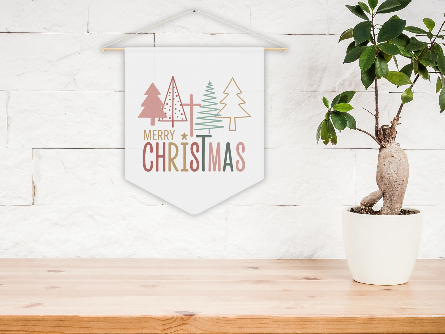 Decorative Christmas wall banner with trees and 'Merry Christmas' text on a white wall above a wooden surface with a plant.