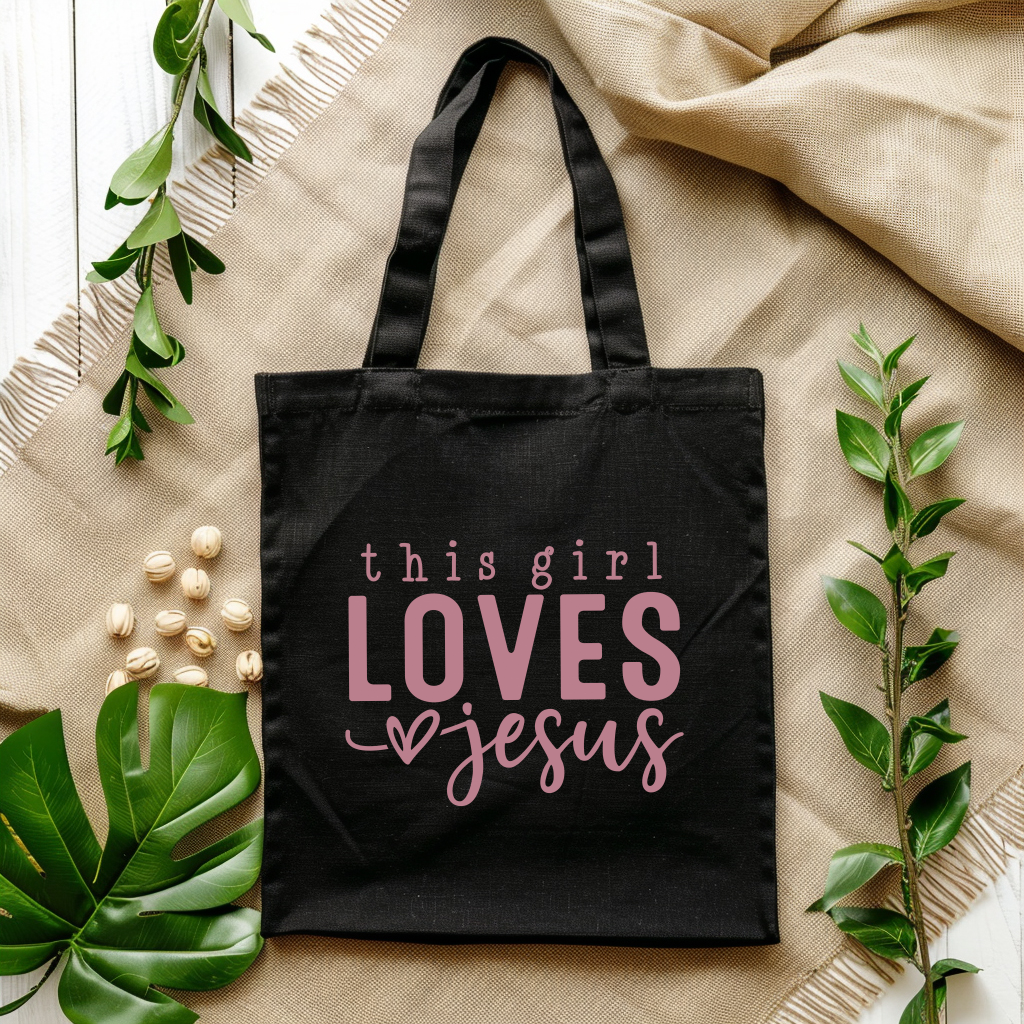 Black tote bag with 'this girl loves Jesus' text on a beige fabric background with green leaves.