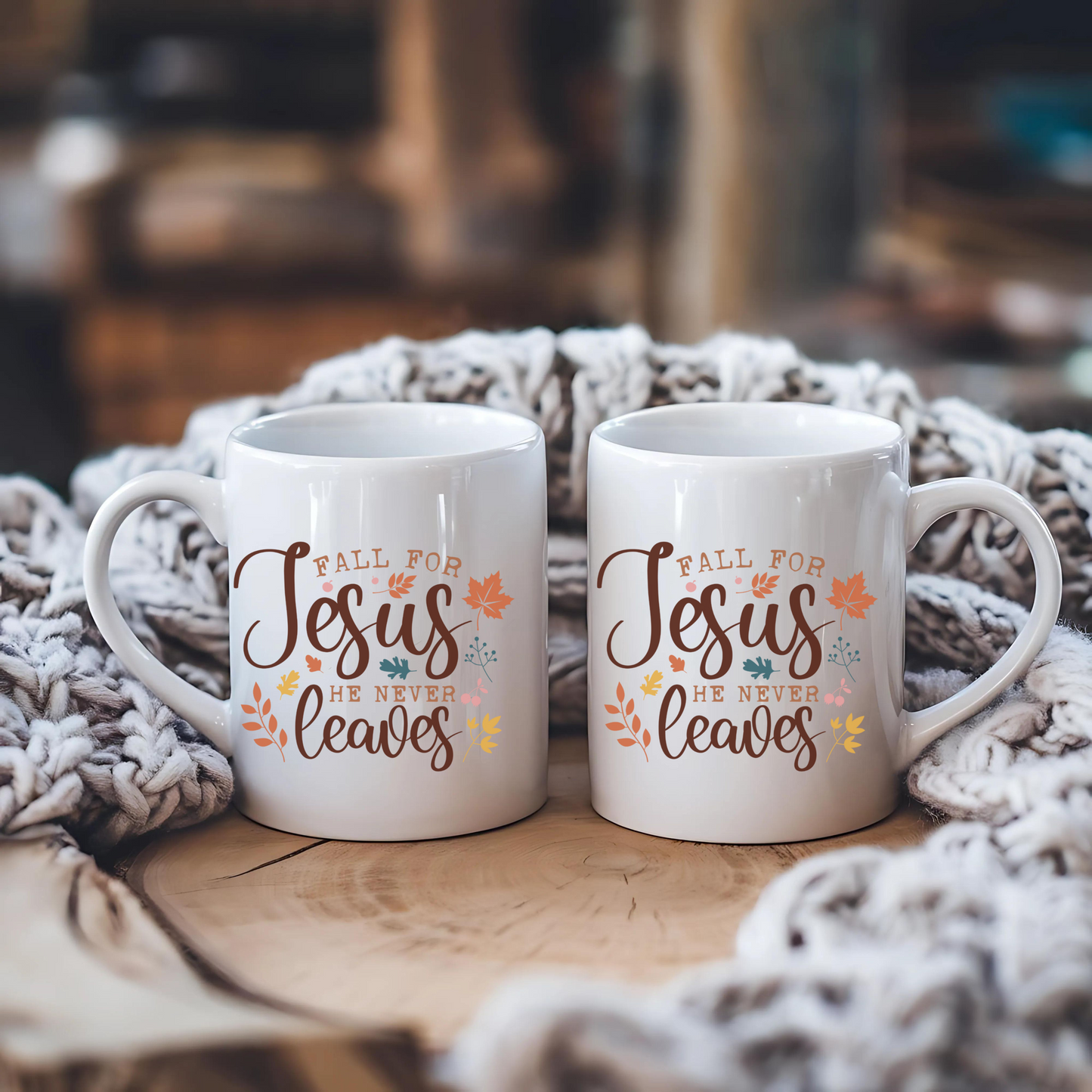Two white mugs with 'Fall for Jesus He Never Leaves' text on a wooden surface with a textured blanket.