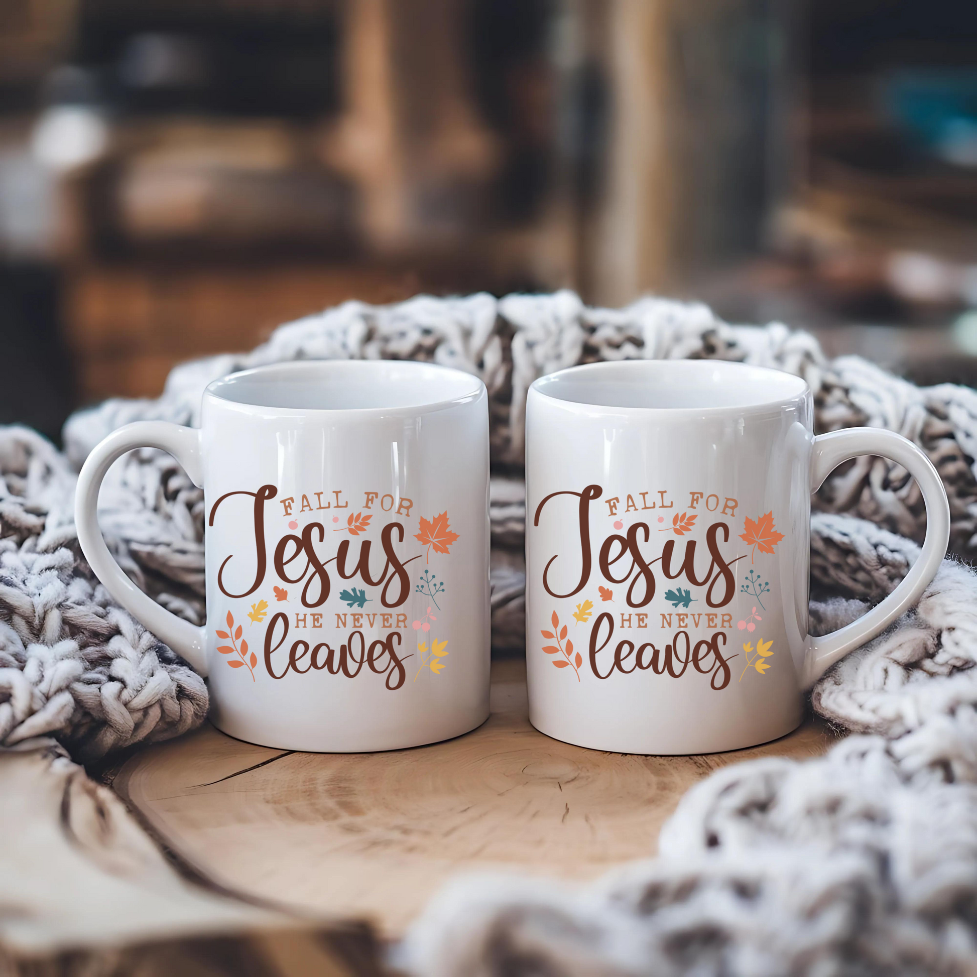 Two white mugs with 'Fall for Jesus He Never Leaves' text on a wooden surface with a textured blanket.