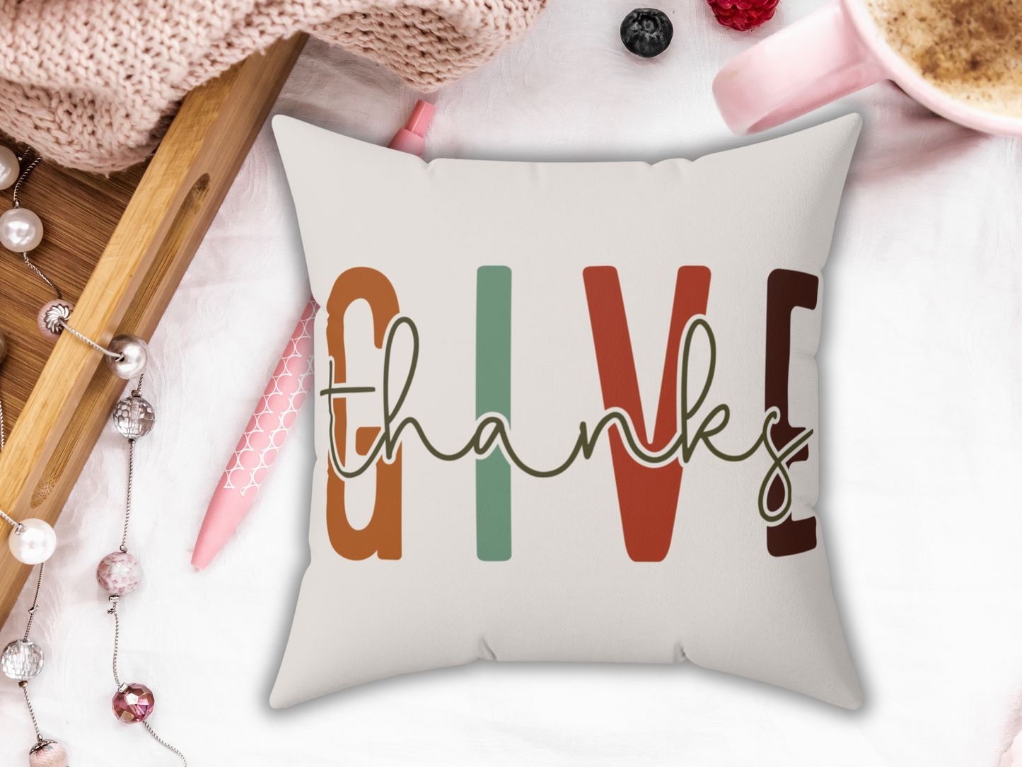 A cozy scene showing a decorative pillow with the words "GIVE thanks" in autumn colors. Nearby are a pink pen, cream, and festive ornaments, evoking warmth.