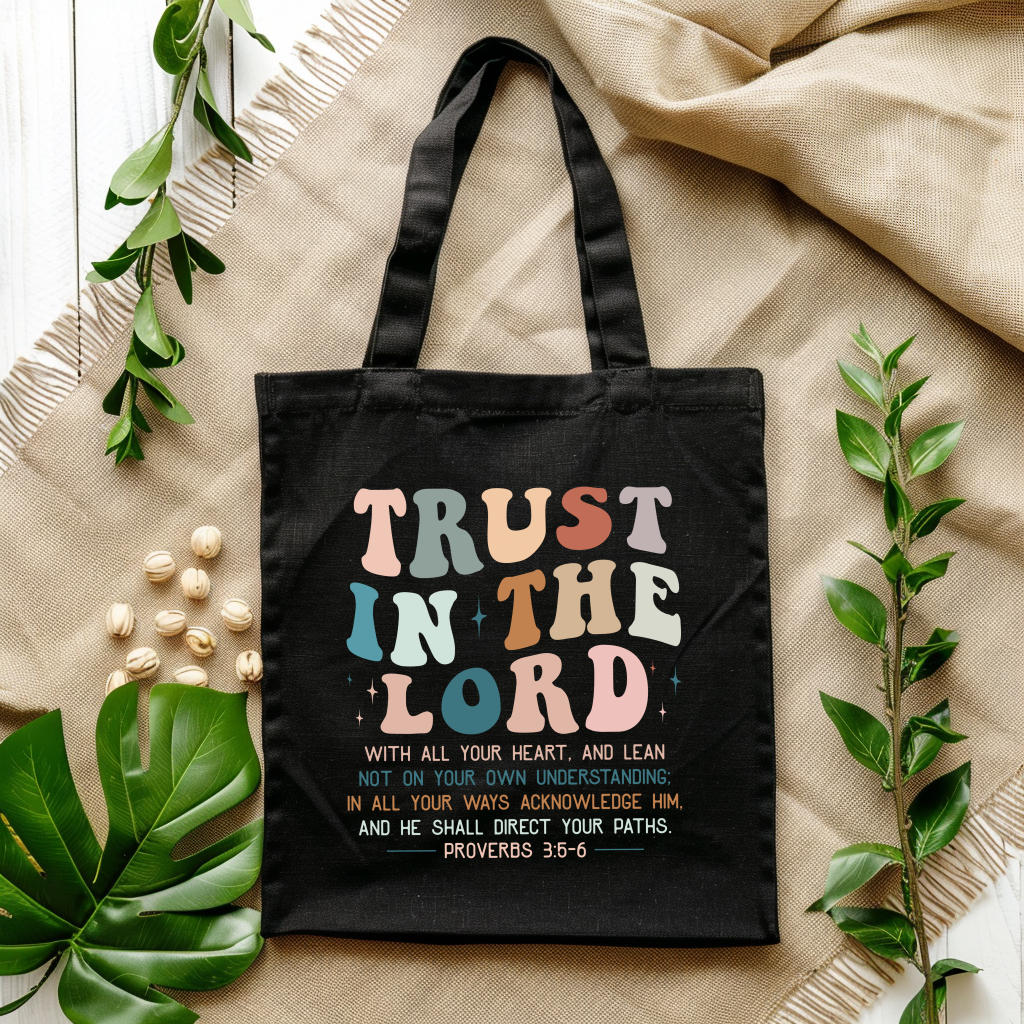 Black tote bag with colorful text 'Trust in the Lord' on a beige fabric background with green leaves.