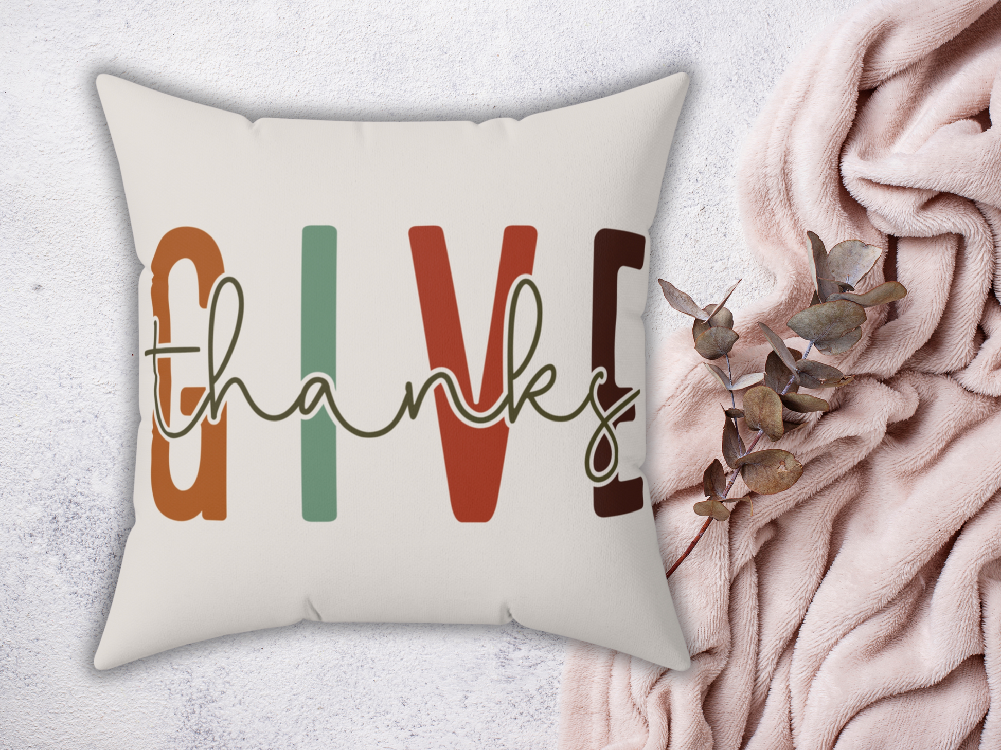 Cushion with "Give thanks" text in colorful letters on a light background. Nearby, a soft pink fabric and eucalyptus branch add a cozy feel.