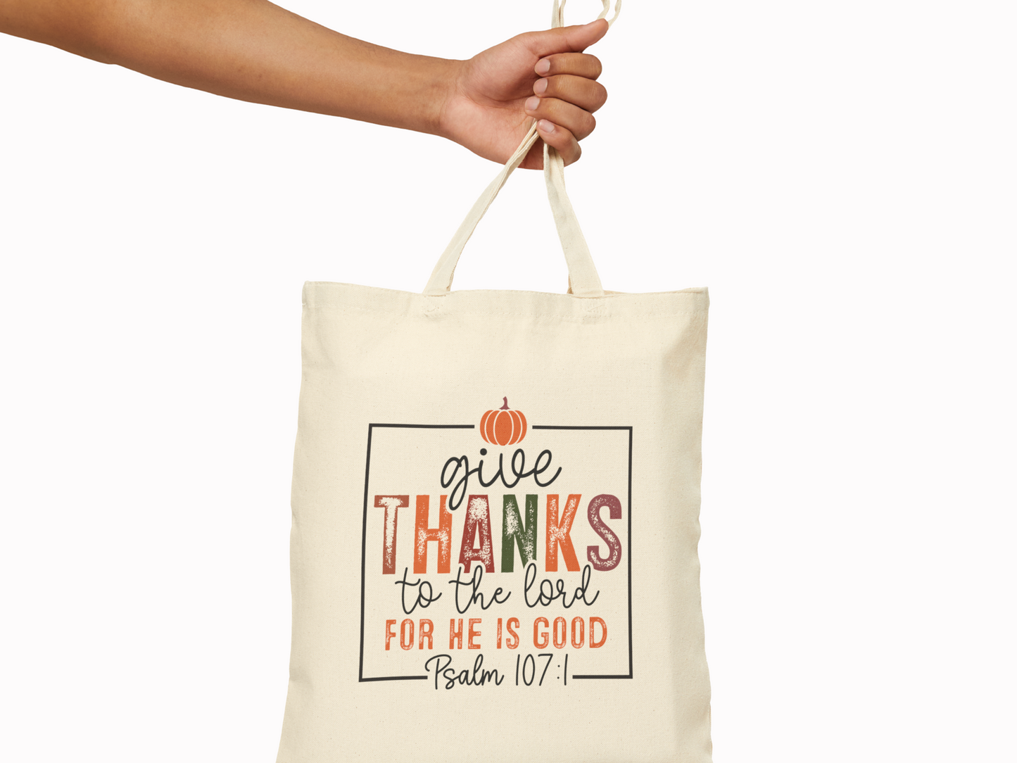 A hand holds a beige tote bag with a quote in decorative fonts. It reads "Give thanks to the Lord for He is good, Psalm 107:1" with a small pumpkin above. The tone is thankful and autumnal.