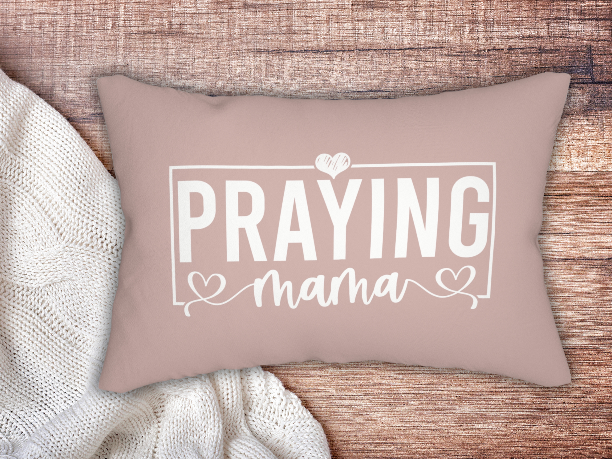 Blush pink scripture pillow with “Praying Mama” text in white, styled on wooden surface beside a cozy cream knit blanket.