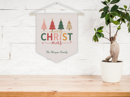 Decorative Christmas wall pennant with colorful trees and text, hanging on a wall above a wooden surface with a plant.
