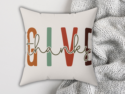 Decorative pillow featuring the words "give thanks" in bold, colorful letters. It's placed on a cozy, textured gray knit blanket, conveying warmth.