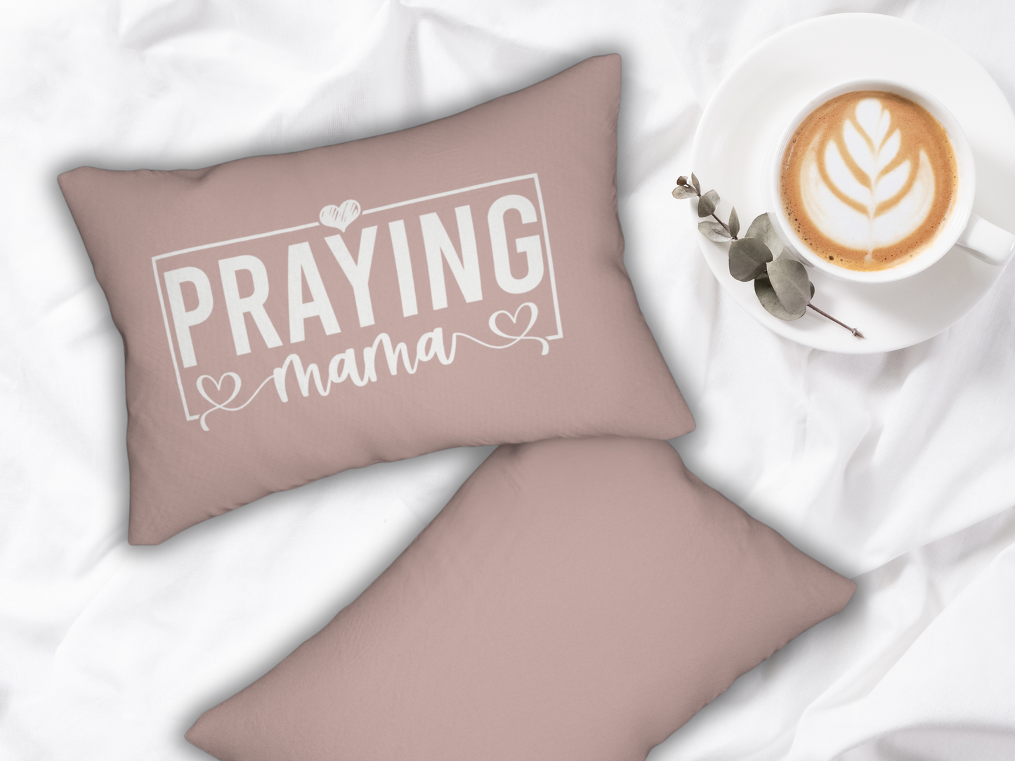 Pair of blush pink “Praying Mama” pillows styled on white bedding beside a latte in a white cup with leaf latte art and eucalyptus sprig.