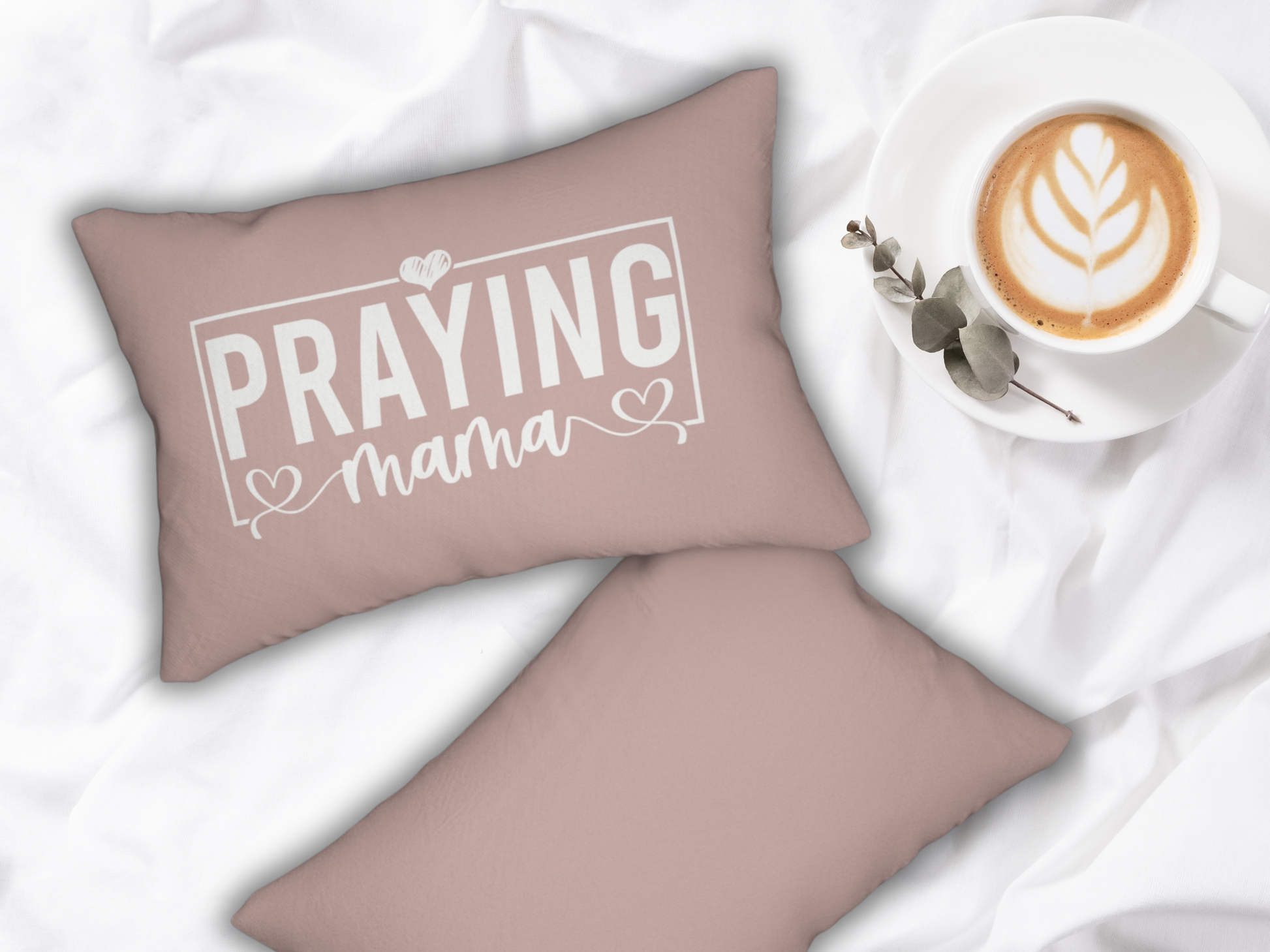 Pair of blush pink “Praying Mama” pillows styled on white bedding beside a latte in a white cup with leaf latte art and eucalyptus sprig.